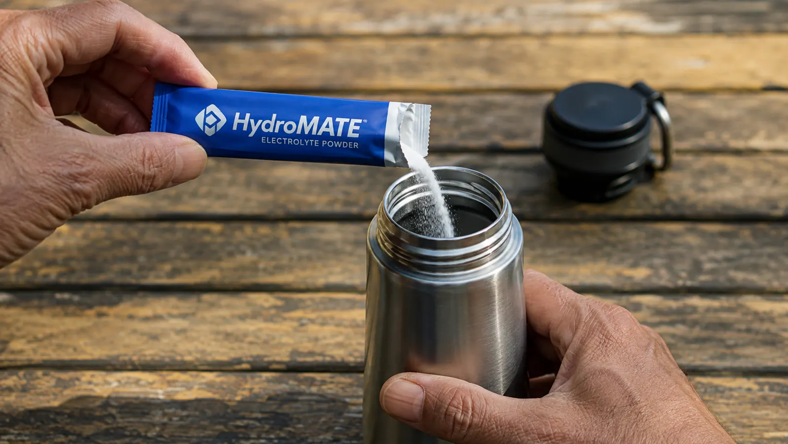 Hand tearing open a HydroMATE electrolyte packet over an open water bottle on a camp table
