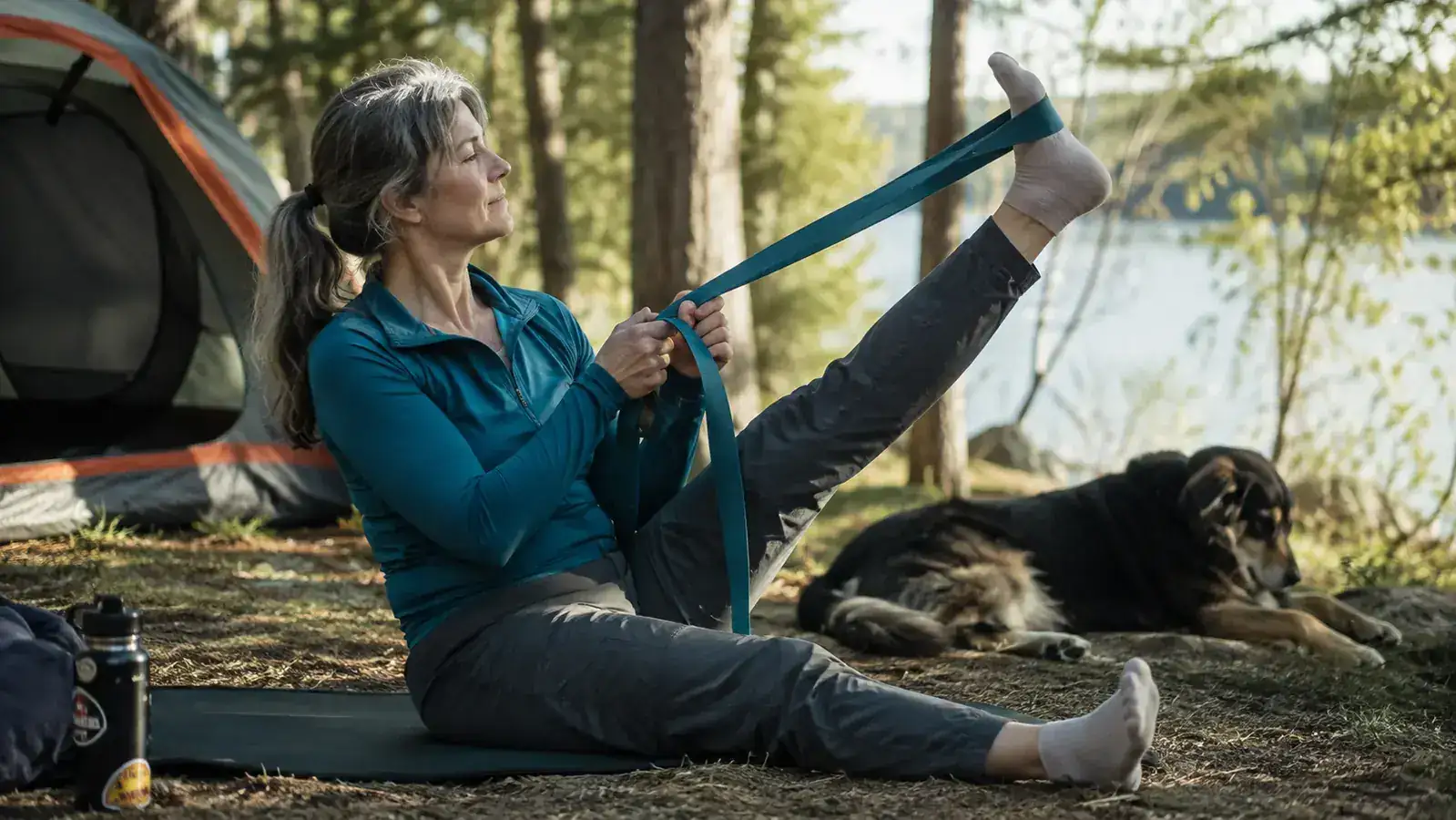 Woman in her 50s stretching her hamstring with a strap outside a tent at sunrise