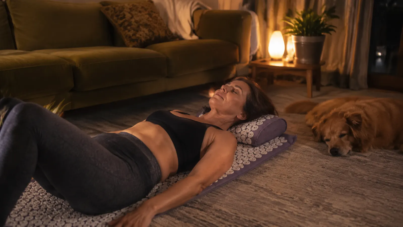 Woman relaxing on an acupressure mat in a cozy living room in the evening with a dog resting nearby