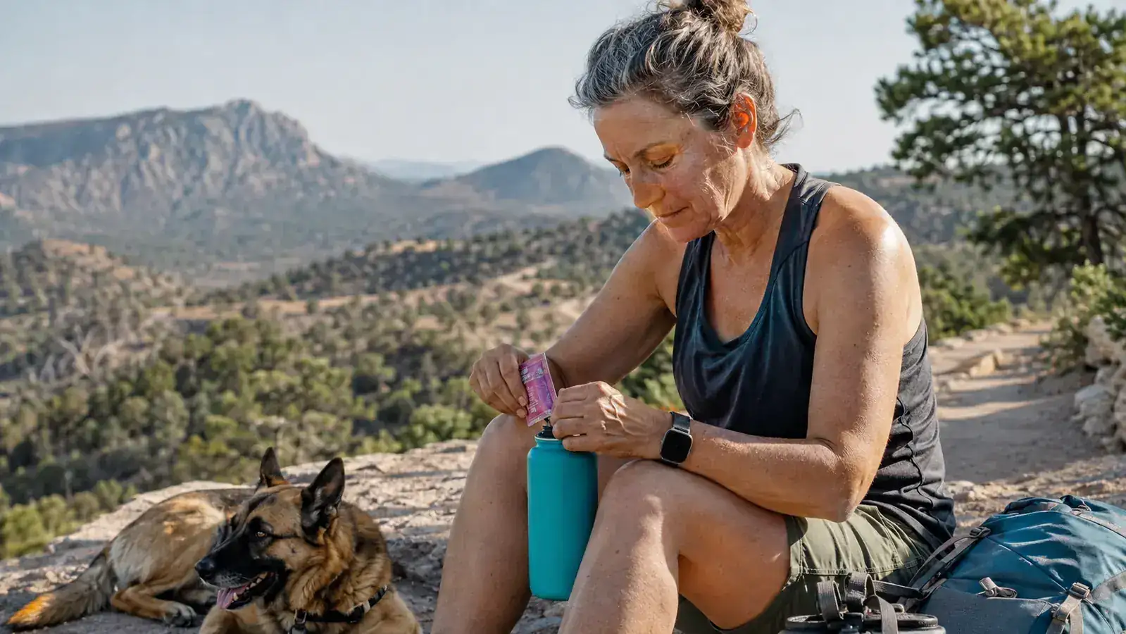 Woman in her early fifties resting on a boulder mid-hike, tearing open an electrolyte packet next to a water bottle, dog panting beside her on the trail