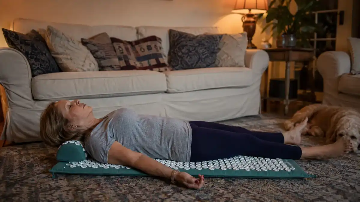 Woman in her 50s lying on her back on a spiky acupressure mat on a living room floor in the evening, shoulders and upper back resting on the mat, small pillow under her neck