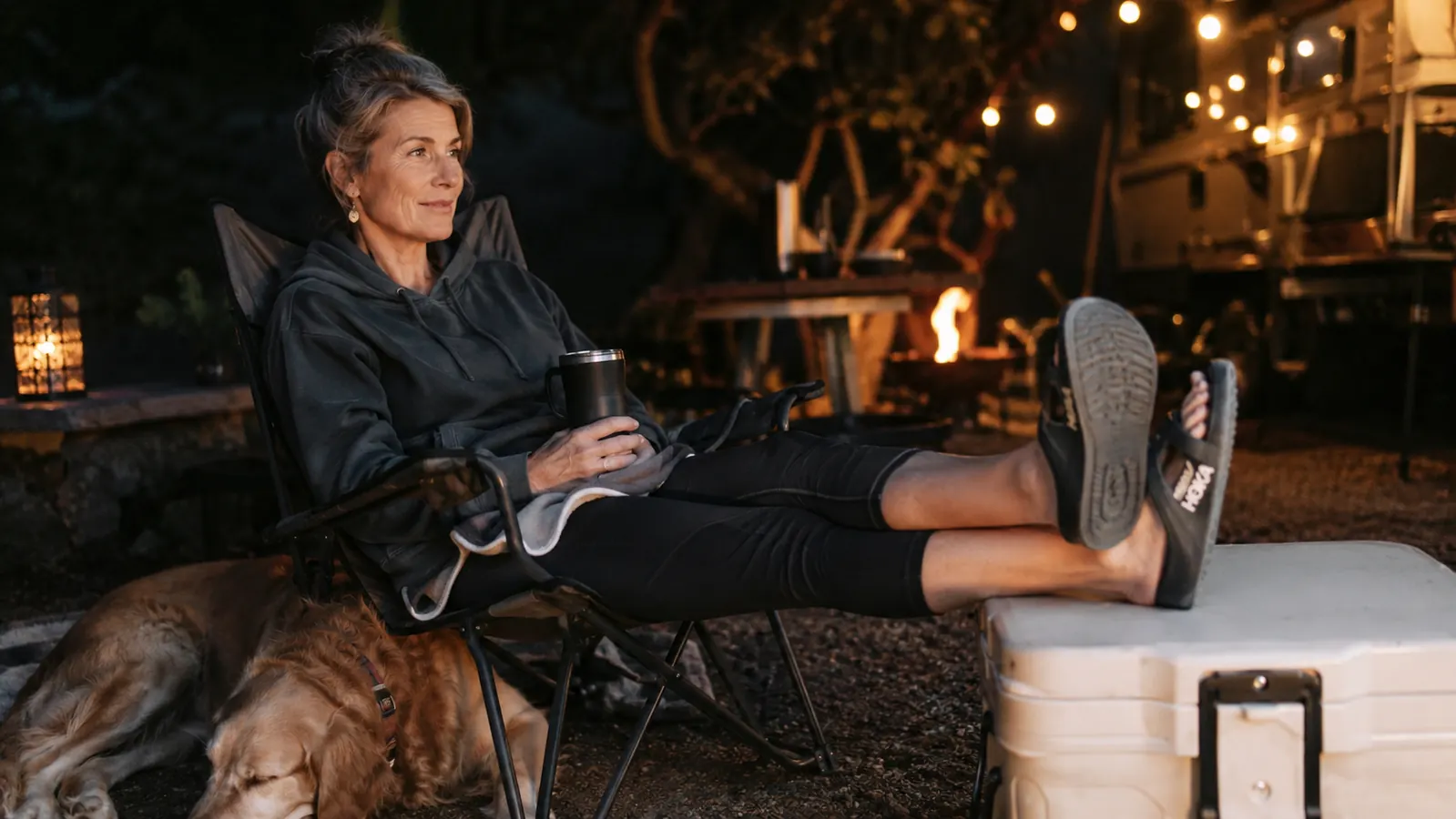 Woman relaxing in a camp chair with her feet up wearing recovery slides, dog resting beside the chair at dusk