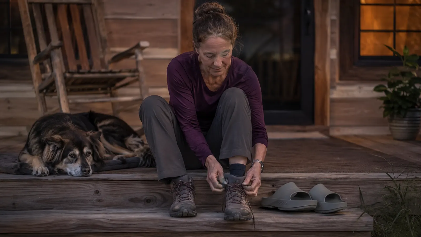 Woman in her early fifties sitting on a back porch step at dusk, taking off a hiking boot with a pair of gray OOFOS recovery slides waiting beside her, dog resting nearby