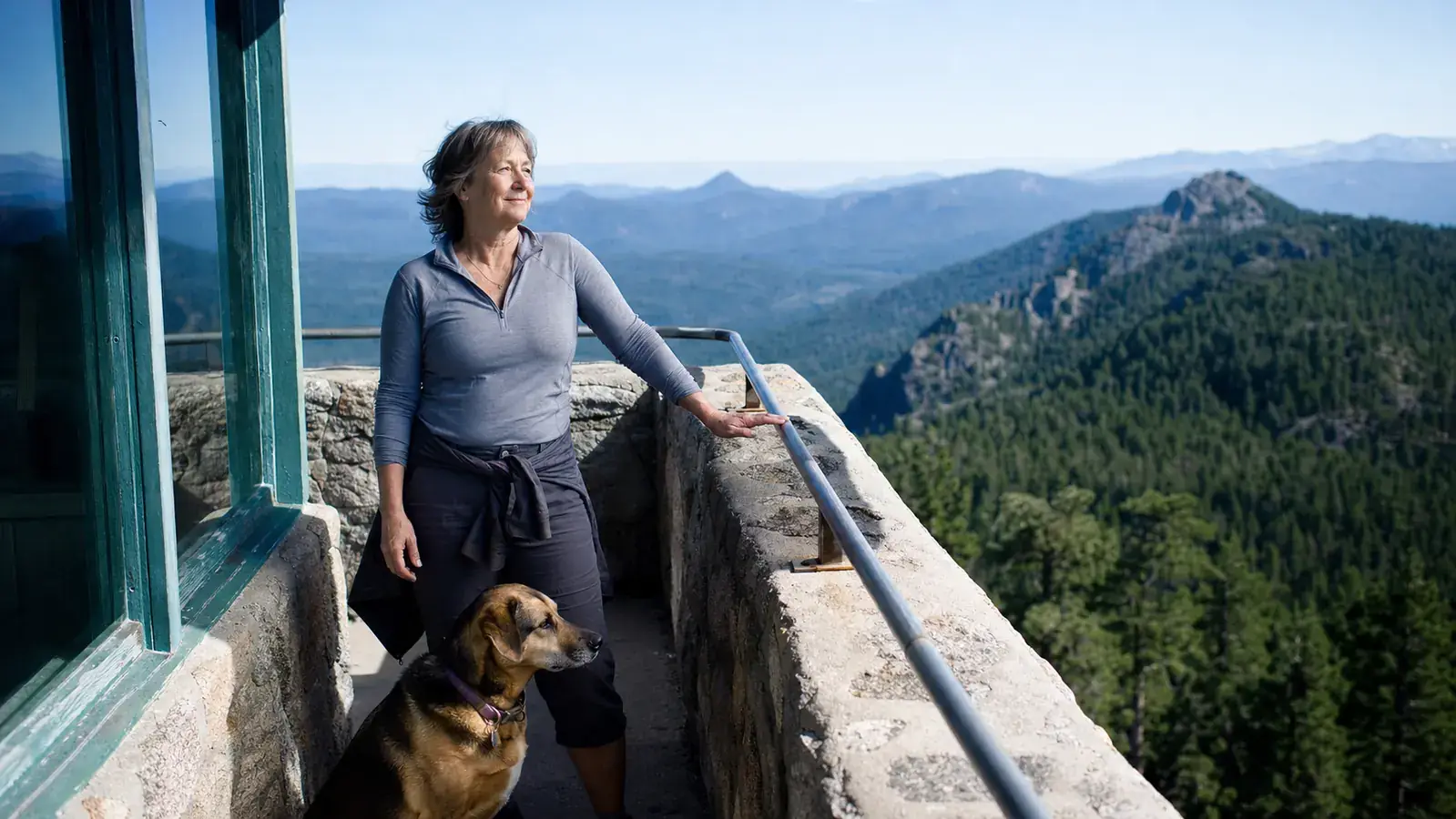Woman and heeler mix dog standing at a granite fire lookout tower overlooking a forested ridge
