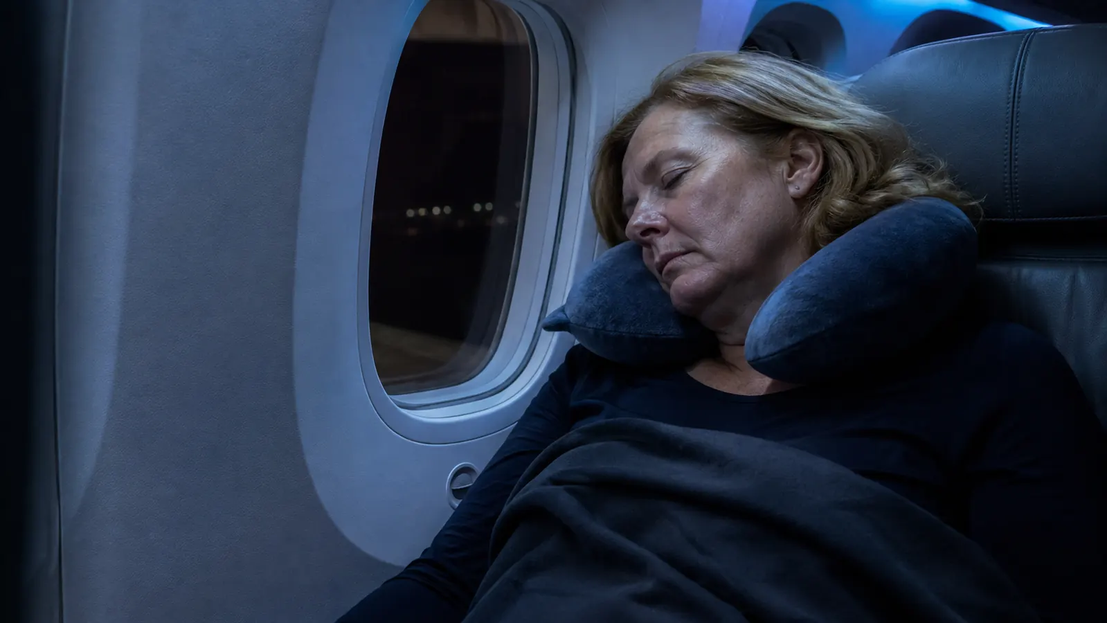 Woman resting her head against an airplane window wearing a gray memory foam travel neck pillow, cabin lights dimmed